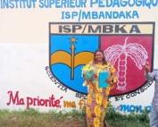 Eine Frau in einem edlen bunten Kleid steht vor der pädagogischen Hochschule in Mbandaka. Auf der Wand hinter ihr ist das Logo der Hochschule zu sehen. Die Frau hält in ihren Händen einen Blumenstrauß und ihre Bachelorarbeit.