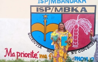 Eine Frau in einem edlen bunten Kleid steht vor der pädagogischen Hochschule in Mbandaka. Auf der Wand hinter ihr ist das Logo der Hochschule zu sehen. Die Frau hält in ihren Händen einen Blumenstrauß und ihre Bachelorarbeit.